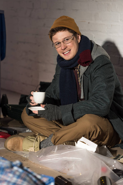 sonriente hombre sin hogar en vasos beber café mientras está sentado en basurero
 - Foto, Imagen