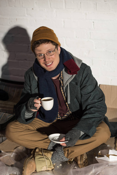 dakloze man drinken koffie zittend door witte bakstenen muur omgeven door vuil glimlachen - Foto, afbeelding