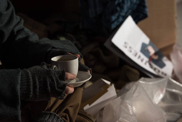 selective focus of homeless man holding coffee cup while sitting on rubbish dump - Photo, image