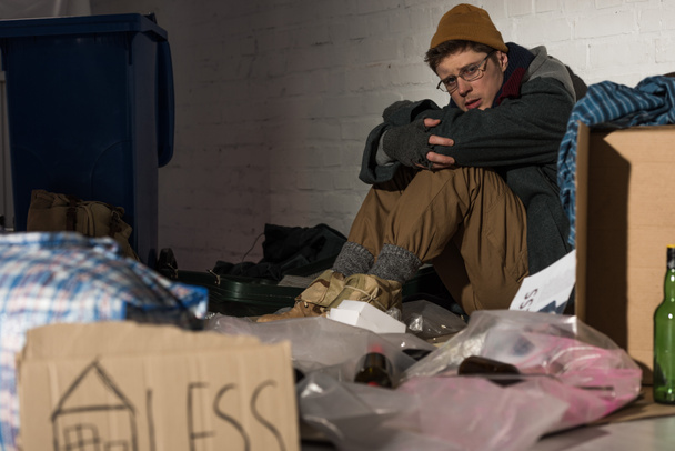 selective focus of depresses homeless man sitting by brick wall on rubbish dump - Φωτογραφία, εικόνα