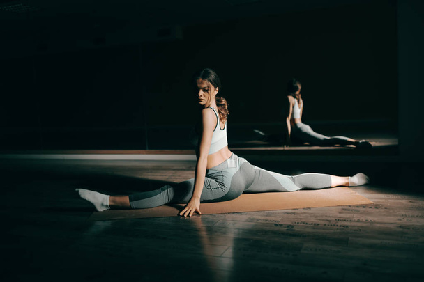 Jonge Kaukasische flexibele brunette in sportkleding beoefening op de mat met gespreide benen. In achtergrond spiegel. - Foto, afbeelding