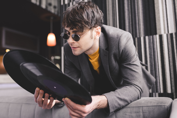 стильный молодой человек в солнцезащитных очках смотрит на ретро-виниловые пластинки
 - Фото, изображение
