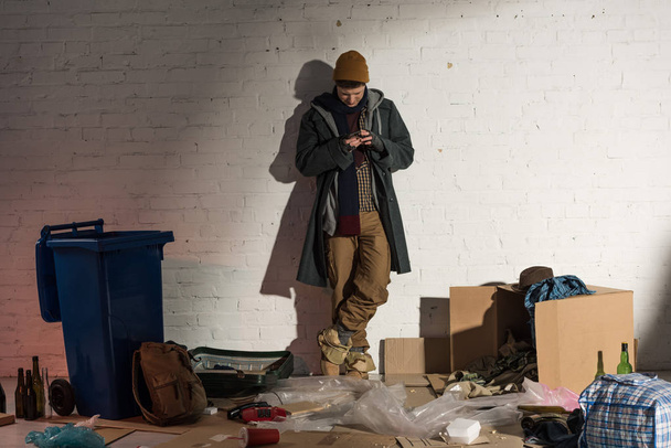 homeless man using smartphone while standing by white brick wall surrounded by rubbish - Fotografie, Obrázek