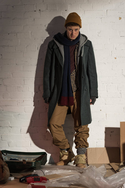 sad homeless man standing by white brick wall and looking at camera - Photo, Image