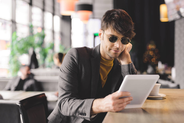 onnellinen nuori mies aurinkolaseissa istuu baaritiskillä ja pitää digitaalista tablettia kupin lähellä kahvilla
 - Valokuva, kuva