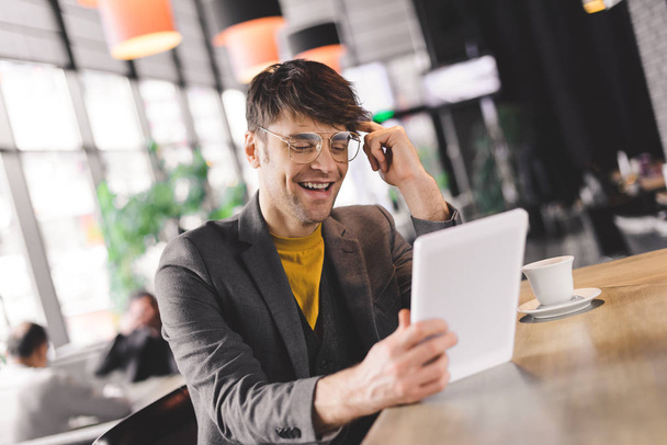 uśmiechający się młody człowiek w okularach siedzi licznik bar patrząc na cyfrowego tabletu w pobliżu kubek z kawą - Zdjęcie, obraz