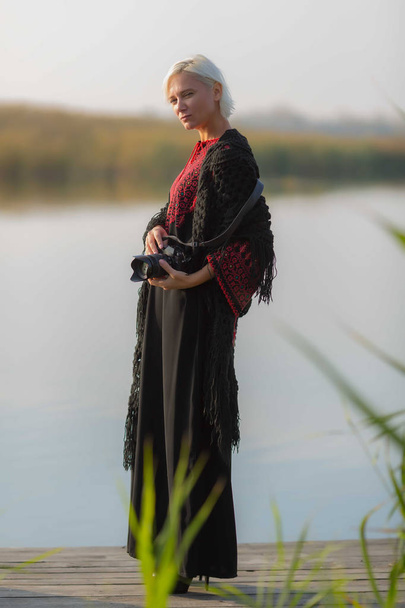 En la primavera, una hermosa mujer en un vestido bordado de color burdeos descansa en un muelle de madera cerca del agua tranquila con una cámara en buen tiempo
. - Foto, imagen