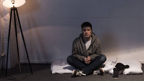 homem sentado no colchão branco dispostos no chão no quarto escuro
 - Foto, Imagem