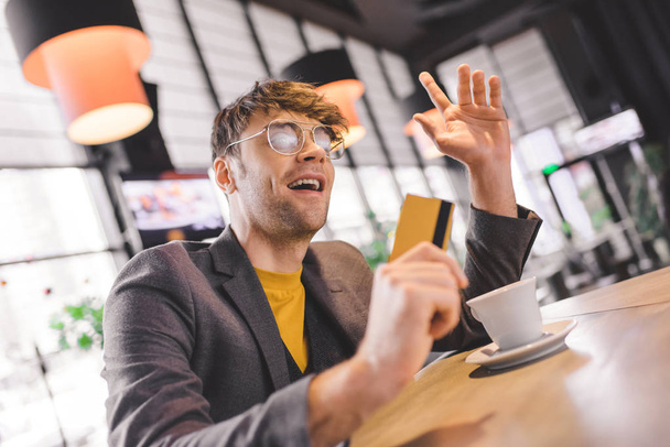 handsome man in glasses gesturing at bar counter while holding credit card near cup with coffee - Photo, Image