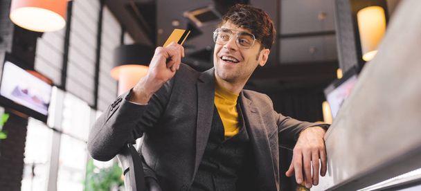 homem feliz em óculos sentado no balcão do bar enquanto segurando cartão de crédito no café
 - Foto, Imagem