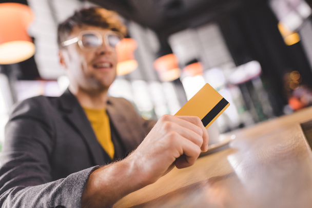 selective focus of credit card in hand of cheerful man in glasses at bar counter - Photo, image