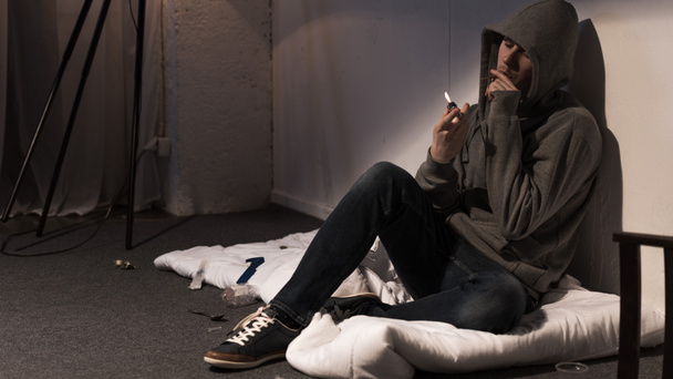 narkoman człowiek palenia marihuany walcowane, papieros biały siedzi na materacu na podłodze - Zdjęcie, obraz