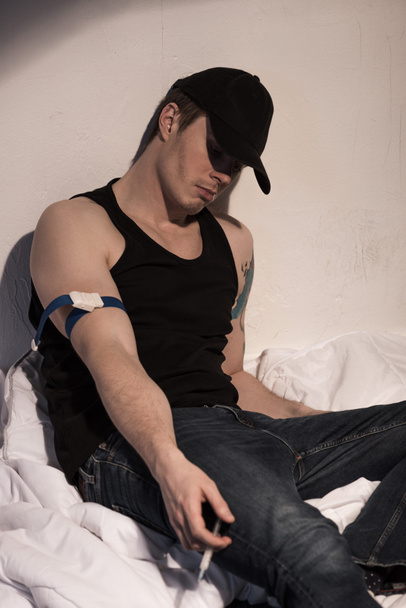 adicto hombre sosteniendo jeringa con dosis de heroína mientras está sentado en la cama
 - Foto, Imagen