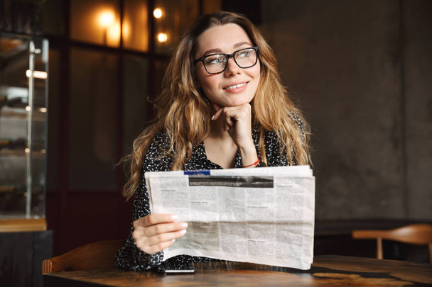 Image of a beautiful young woman sitting in cafe indoors reading newspaper. - Photo, image