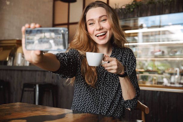 Kuva emotionaalinen onnellinen kaunis nuori kaunis nainen istuu kahvilassa sisällä matkapuhelimella puhuminen ystävien kanssa ottaa selfie juo kahvia
. - Valokuva, kuva
