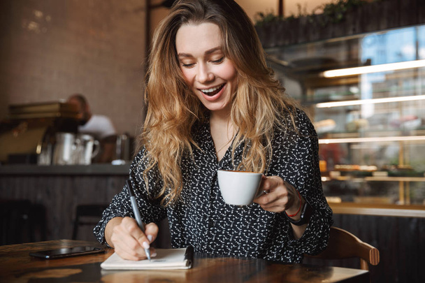 Photo of beautiful young pretty woman sitting in cafe indoors writing notes in notebook drinking coffee. - Fotoğraf, Görsel