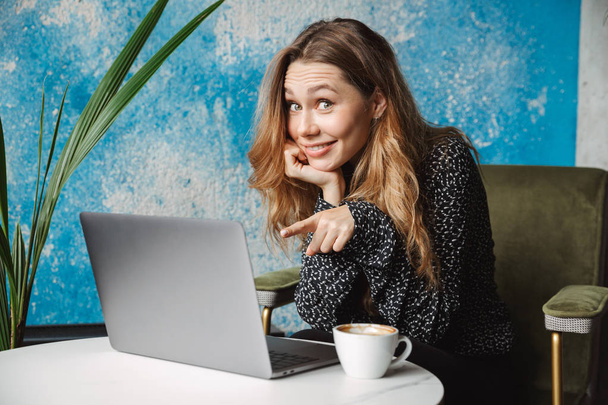 Foto van gelukkig emotionele mooie jonge mooie vrouw zitten in Cafe binnenshuis met behulp van laptopcomputer koffie drinken. - Foto, afbeelding