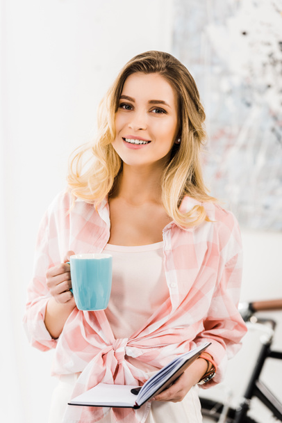 Attractive blonde woman in checkered shirt holding coffee cup and notebook - Foto, Imagem