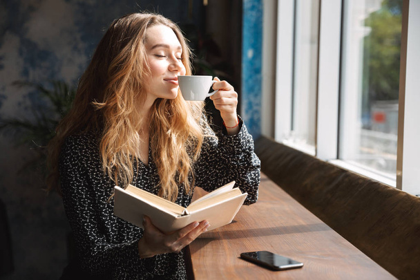 Beautiful young woman sitting at the cafe indoors, reading book, drinking coffee - Foto, Imagem