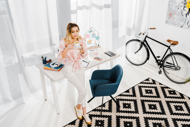 Mujer rubia bonita en pantalones blancos bebiendo café en casa oficina
 - Foto, imagen