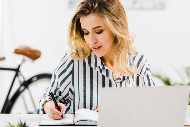 Charming blonde woman in striped shirt writing in notebook at workplace - Fotoğraf, Görsel