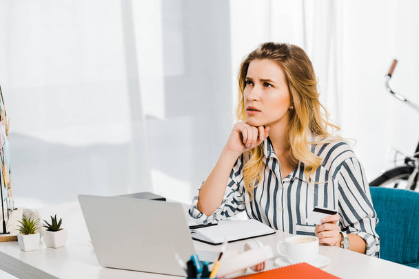 Pensive woman in striped shirt holding credit card at workplace - Foto, Imagen