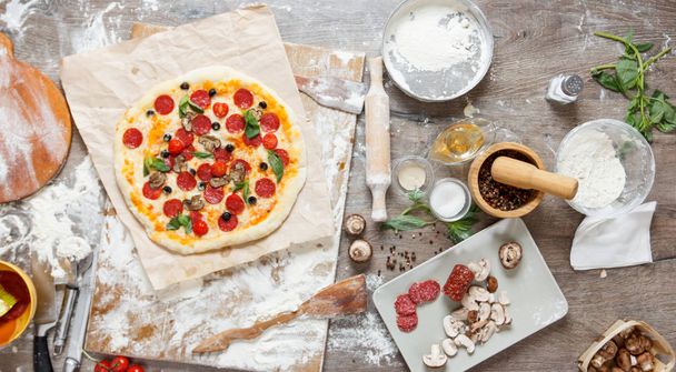 Top overhead view cooking pizza with ingredients, tomatoes, salami, flour, dough basil, spices, mozzarella and mushrooms on wooden tabletop - Photo, Image