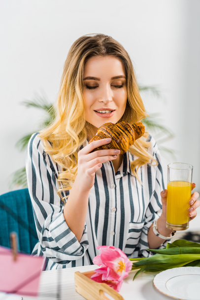 Curly blonde woman holding croissant and drinking orange juice - Valokuva, kuva