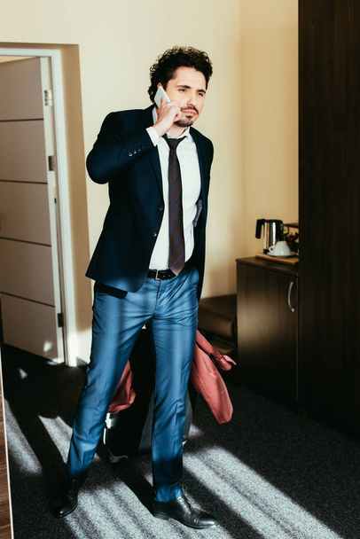 businessman in suit talking on smartphone in hotel room with suitcase - Fotó, kép