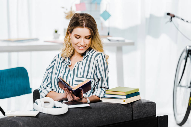 Улыбающаяся девушка в полосатой рубашке сидит на диване и читает книгу
 - Фото, изображение