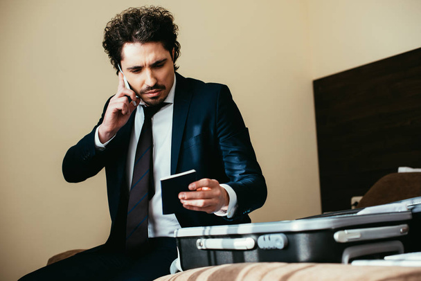 handsome businessman talking on smartphone and holding passport while sitting on bed with luggage in hotel room - Foto, imagen