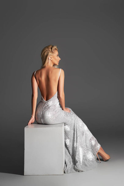 High fashion portrait of young elegant woman. Young lady in amazing silver glitter sequins luxury dress sitting on  armchair Studio shot. Back view  - Foto, Imagen