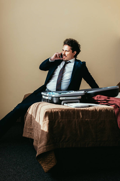businessman in suit talking on smartphone while sitting on bed with suitcase in hotel room - Photo, Image