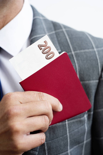 closeup of a young caucasian businessman, wearing an elegant gray suit, taking in or taking off his passport, with a 200 hundred banknote in it, from the top pocket of his jacket - Fotó, kép