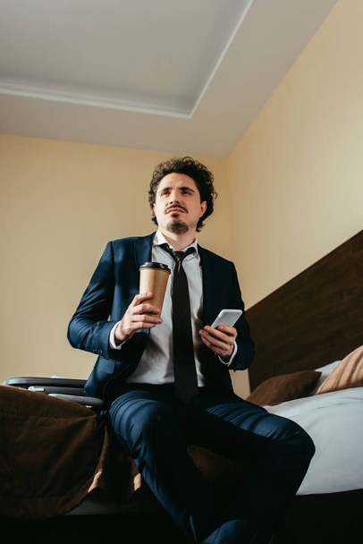 thoughtful businessman using smartphone and holding coffee to go in hotel room - Foto, Imagem