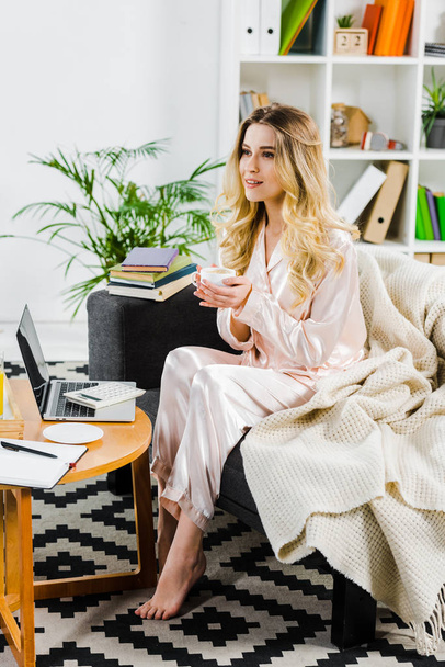 Relaxed woman in pyjamas sitting on sofa and drinking coffee in morning - Photo, Image