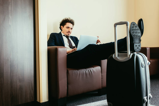 hombre de negocios teletrabajo en el ordenador portátil con las piernas en la bolsa de viaje en la habitación de hotel
 - Foto, imagen