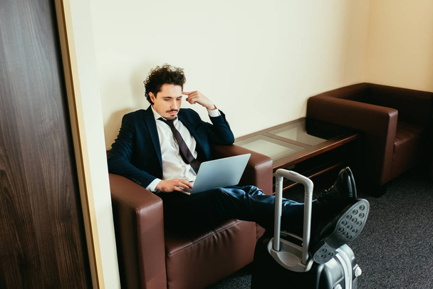 hombre de negocios que trabaja en el ordenador portátil con las piernas en la maleta en la habitación del hotel
 - Foto, Imagen