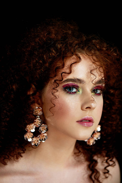 A girl with lush curly red hair. Close face with bright make-up and massive earrings. Purple shadows. - Φωτογραφία, εικόνα