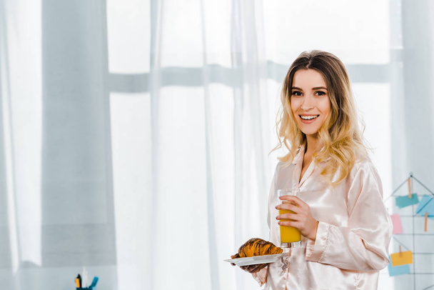 Laughing young woman in pyjamas holding croissant and glass of orange juice - Foto, Bild