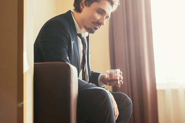pensive businessman in formal wear holding glass of cognac in hotel room  - Foto, Bild