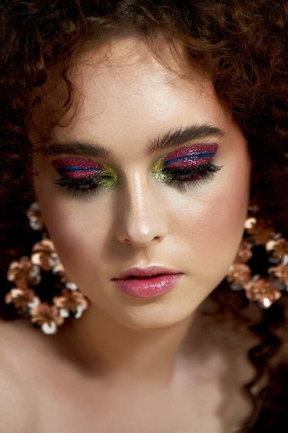 A girl with lush curly red hair. Close face with bright make-up and massive earrings. Purple shadows. - Foto, afbeelding