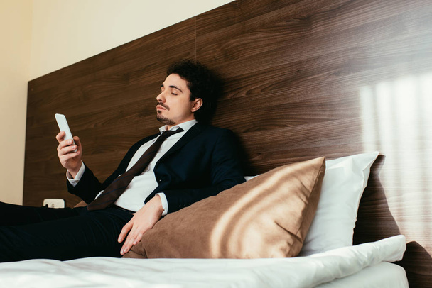 businessman in suit using smartphone on bed in hotel room - Zdjęcie, obraz