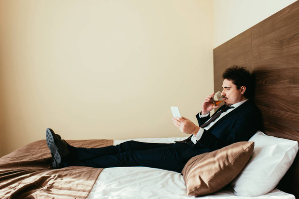 businessman in formal wear using smartphone and drinking whiskey on bed in hotel room - Foto, Bild