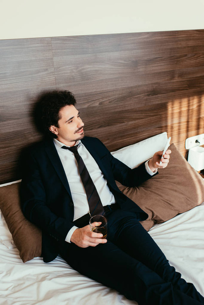 бізнесмен використовує смартфон і тримає келих коньяку на ліжку в готельному номері
 - Фото, зображення