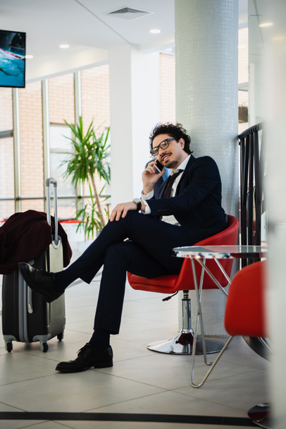 sonriente hombre de negocios hablando en el teléfono inteligente mientras espera en el vestíbulo del hotel con bolsa de viaje
 - Foto, Imagen