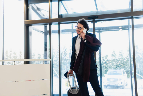smiling businessman with passport and luggage talking on smartphone in hotel lobby - 写真・画像