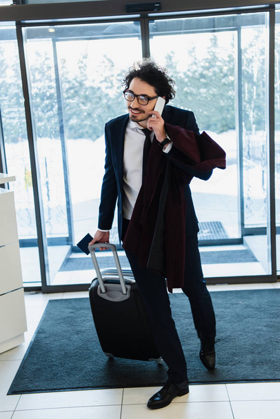 homme d'affaires souriant avec passeport et sac de voyage parlant sur smartphone dans le hall de l'hôtel
 - Photo, image