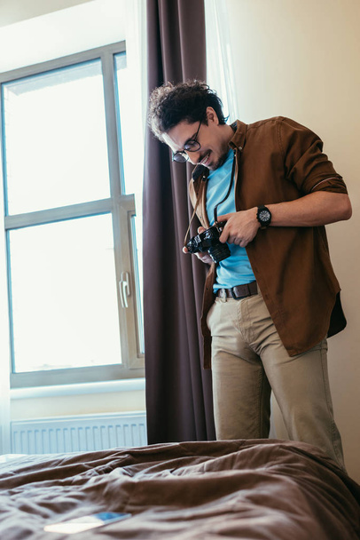 man in eyeglasses looking at photo camera near window in hotel room - Fotoğraf, Görsel