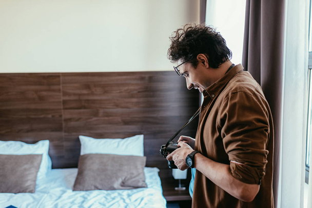 male photographer in eyeglasses looking at photo camera in hotel room - Fotó, kép